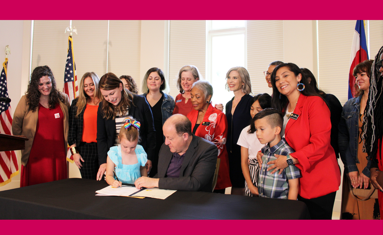 A young girl stands next to Colorado Gov. Jared Polis and is surrounded by women wearing red as he signs the 2023 Ensure Equal Pay for Equal Work Act.