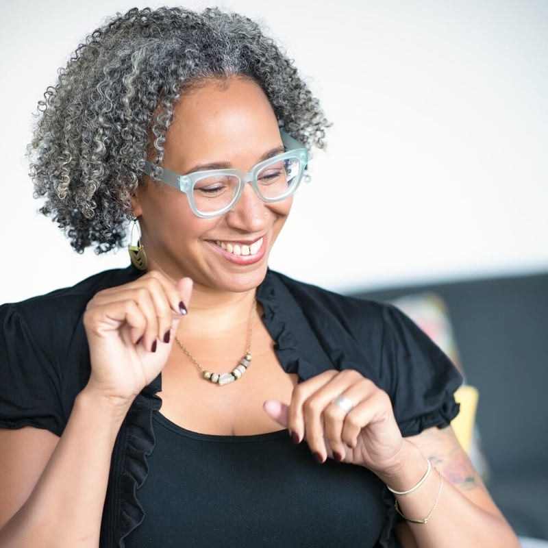 Mia Birdsong - a Black woman with gray curly hair, glasses, and short sleeved black shirt, looks down while smiling