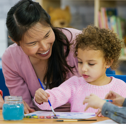 Early child care worker looking over young child painting